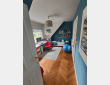 Ein modernes Kinder- oder Arbeitszimmer mit blauen Wänden, einem Schreibtisch samt Computer und rotem Bürostuhl, einem Regal mit Spielsachen und Büchern sowie einem blauen Gymnastikball. Der Boden ist aus Holz in Fischgrätmuster, und ein großes Fenster mit Jalousien lässt Tageslicht herein.