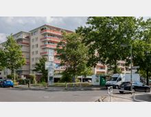 Mehrstöckiges Wohngebäude mit hellbeiger Fassade und orangen Balkonen, umgeben von Bäumen. Im Vordergrund befindet sich eine Straßenkreuzung mit geparkten Autos, einem Müllcontainer und Wegweisern.