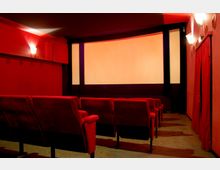 Kleiner Kinosaal mit roten Wänden und roten gepolsterten Kinosesseln, die auf eine große, unbeleuchtete Leinwand ausgerichtet sind. An den Wänden befinden sich Wandlampen und rechts hängt ein roter Vorhang.