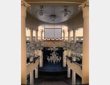 Innenraum eines Veranstaltungsortes mit ovaler Grundstruktur, hohen weißen Säulen und mehreren gläsernen Kronleuchtern. Im Hintergrund sind eine Projektionsfläche und eine Bühne sichtbar, die von speziell montierten Beleuchtungselementen umgeben sind.