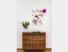 Holzkommode mit schlichter, geradliniger Optik vor einer weißen Wand; darüber ein Organizer mit mehreren weißen Fächern, die mit Alltagsgegenständen wie einem Pass, Schreibutensilien und Schlüsseln gefüllt sind. Auf der Kommode steht eine Vase mit bunten Blumen und daneben ein kleines Tablett mit Brille und weiteren Utensilien.