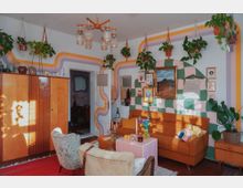 Wohnzimmer mit kreativer Retro-Einrichtung: An den Wänden befinden sich farbige, wellenförmige Muster in Orange, Lila, Grün und Rosa, ergänzt durch hängende Pflanzen. Ein orangefarbenes Sofa, ein rosa Kacheltisch, Vintage-Möbel aus Holz und dekorative Bilder und Makramee-Wandbehänge prägen den Raum, der von warmem Tageslicht erleuchtet wird.