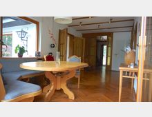 Innenraum eines Wohnzimmers mit Holzboden und Holzdecke, einem runden Esstisch mit Eckbank und Stühlen sowie einer Glastür im Hintergrund. Links steht eine große Fensterfront mit Pflanzen, und rechts ein kleiner Holztisch mit Dekoration.