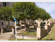 Terrassierter Garten vor einem historischen Gebäude mit symmetrisch gepflanzten Bäumen und dekorativen Steingeländern. Große Blumentöpfe mit Pflanzen zieren die Geländer, und ein blauer Papagei aus Kunststoff ist auf einem weißen Pfosten befestigt.