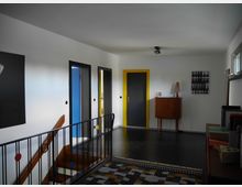 Flur in einem modernen Wohnhaus mit weißen Wänden, einer schwarzen Treppe mit bunten Kreisen im Geländer und Türen mit gelben Rahmen. Im Hintergrund steht eine hölzerne Kommode mit einer Tischlampe, daneben hängt ein Bild mit mehreren Schwarz-Weiß-Fotografien.