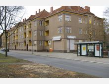 Wohngebäude aus der frühen 20. Hälfte des 20. Jahrhunderts mit brauner Putzfassade und gelben Fensterrahmen sowie Balkonen, daneben ein moderner Bushaltestellenunterstand mit Glaswänden und Sitzbank an einer ruhigen städtischen Straße. Im Erdgeschoss befinden sich geschlossene Ladenfronten mit Rollläden.