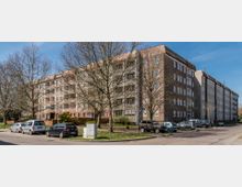 Mehrstöckiges Wohngebäude aus Backstein mit Balkonen; davor stehen mehrere Autos auf Parkplätzen entlang einer Straße, umgeben von kahlen Bäumen. Der Himmel ist klar und blau.