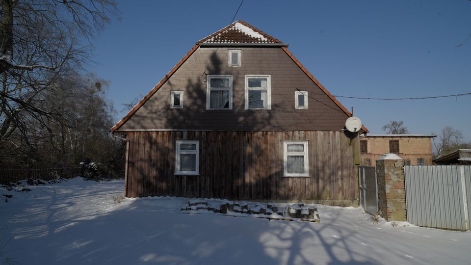 Ein freistehendes, älteres Wohnhaus mit steilem Satteldach und Holzfassade steht in einer verschneiten Hof- oder Gartenfläche. Mehrere kleine Fenster, eine Satellitenschüssel an der rechten Seite sowie angrenzende Zäune und Nebengebäude sind vor blauem Winterhimmel zu sehen.