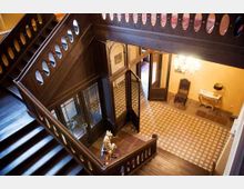 Großes Treppenhaus eines historischen Gebäudes mit dunklem Holzgeländer und Balustraden. Der Boden im Eingangsbereich ist mit gemusterten Fliesen gestaltet, und es gibt eine dekorative Beleuchtung sowie antike Möbel an den Wänden.