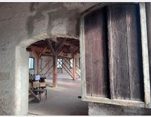 Ein Blick in einen historischen Innenraum mit unverputzten Steinwänden, hölzernen Trägerbalken und einer großen geöffneten Holztür. Im Hintergrund sind Fenster mit Rundbögen und einige Stühle zu sehen.