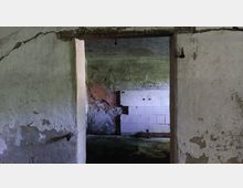 Blick durch eine Türöffnung in einen verfallenen Raum mit beschädigten Wänden, freiliegenden Putzschichten und teils weißen Fliesen. Der Boden ist uneben, und die Wände zeigen Spuren von Feuchtigkeit und Abnutzung.