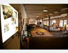 Weiträumiger, loftartiger Innenraum mit einer offenen Struktur, Holzbalkendecke und mehreren Säulen. Entlang der Wände sind Dekorationen, Fotos und Möbelstücke wie Sessel, Pflanzen und Regale zu sehen; in der Mitte liegt ein großer Teppich, während rechts ein Tisch und Stühle arrangiert sind.