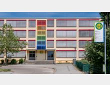 Gelb-beiges mehrstöckiges Gebäude mit geschlossenen Rollläden und bunten, farbigen Lamellenstreifen (rot, gelb, grün, blau) in der Mitte der Fassade, daneben ein Haltestellenschild für Busse mit der Aufschrift „Beelitz Platanenring“. Ein Gehweg führt zum Eingang des Gebäudes, umgeben von Bäumen und Sträuchern.