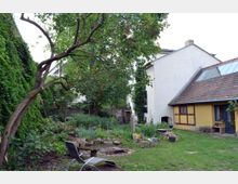 Hinterhof mit Gartenbereich, bestehend aus einer Wiese, Blumenbeeten, einem Feuerplatz und einem großen Baum. Im Hintergrund sind ein gelbes Fachwerkhaus mit Satteldach und ein angrenzendes weißes Wohnhaus zu sehen.