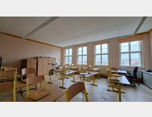 Leeres Klassenzimmer mit hellen Wänden, mehreren Reihen von Schülertischen und hochgestellten Stühlen auf Parkettboden. Große, mehrflügelige Fenster entlang der rechten Wand lassen viel Tageslicht herein; an der Rückwand stehen Schränke und seitlich befindet sich ein Lehrerpult.