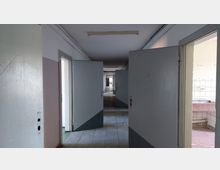 Ein leerer, schmaler Flur in einem Gebäude mit grauen Türen auf beiden Seiten, die teilweise geöffnet sind. Der Boden besteht aus grauen Fliesen, und am Ende des Flurs fällt Tageslicht durch ein Fenster.