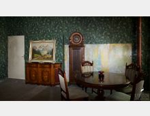 Ein rustikaler Innenraum mit grüner Tapete im Pflanzendesign und einer abgenutzten Wand. Im Vordergrund steht ein runder Holztisch mit Stühlen, daneben eine antike Standuhr und ein Holzbuffet mit einem Landschaftsgemälde darauf.