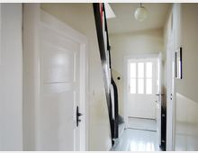 Flur in einem Wohnhaus mit hell gestrichenen Wänden und glänzendem, grauem Holzboden. Ein weißes Holztür-Design mit Glaseinsätzen sowie eine geschwungene, schwarze Treppe dominieren den Raum; an der Decke hängt eine runde Hängeleuchte.