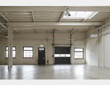 Empty industrial warehouse interior with white-painted brick walls, a polished concrete floor, and exposed beams, ducts, and hanging lights. Several rectangular windows line the wall beside a door and a large black roll-up garage door under a skylight.