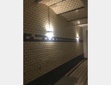 Ein schmaler Flur mit gewölbter Decke und cremefarbenen Ziegelwänden, verziert mit einem horizontalen Streifen aus blauen Fliesen. An den Wänden sind regelmäßig platzierte Wandlampen angebracht, und der Boden ist mit einem Muster aus hellen und dunklen Fliesen gestaltet.