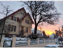 Großes Wohnhaus im traditionellen Stil mit rotem Satteldach und weißen Fensterrahmen, umgeben von verschneiten Bäumen und Gärten. Im Hintergrund ein farbenprächtiger Sonnenuntergang mit orangefarbenem und rosafarbenem Himmel.