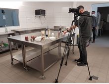 Produktionsküche mit Edelstahlarbeitsflächen, auf denen Flaschen, Schalen und Lebensmittel arrangiert sind. Im Vordergrund steht ein Fotograf mit Kamera auf einem Stativ, während im Hintergrund ein weiterer Mann in Arbeitskleidung zu sehen ist.