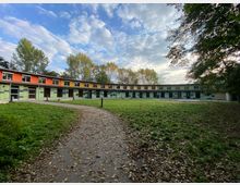 Hufeisenförmige, einstöckige Gebäude mit einer Fassade aus farbigen Paneelen (Orange, Gelb, Grün) umgeben eine zentrale Rasenfläche mit einem gepflasterten Weg. Im Hintergrund stehen hohe, laubtragende Bäume unter einem teilweise bewölkten Himmel.