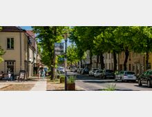 Baumbestandene Stadtstraße mit Gehweg und parkenden Autos entlang niedriger, historischer Gebäude mit Ladenfronten und Cafébestuhlung. In der Straßenmitte stehen eine Laterne mit Banner und ein Schild „Kirchplatz“, die Straße führt geradeaus in ein dicht begrüntes Viertel.