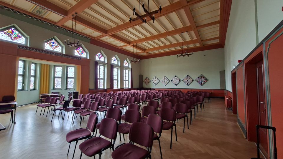 Großer Veranstaltungs- oder Vortragssaal mit Reihen aus bordeauxfarbenen Stühlen auf Holzboden und hohen Fenstern mit Buntglas-Oberlichtern. Sichtbare Holzbalkendecke mit Hängelampen, rote Wandvertäfelung und geometrische Wanddekorationen an der Stirnseite.