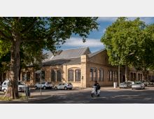Backstein-Gebäude im historischen Stil mit großen Fensterbögen und angeschrägtem Blechdach, umgeben von Bäumen. Im Vordergrund befindet sich eine Straße mit parkenden Autos und einem Rollerfahrer.