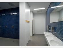 Öffentlicher Dusch- und Waschräum mit blauen Fliesen; links eine blaue geflieste Duschwand mit mehreren Duschköpfen, rechts ein weißes Waschbecken mit drei Becken und Spiegel darüber. Im Hintergrund eine Garderobenleiste und ein Heizkörper an der Wand.