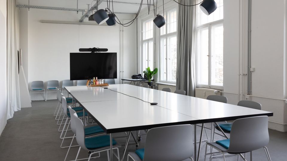 Heller, moderner Besprechungsraum mit langen weißen Konferenztischen und grauen Stühlen, darüber hängende schwarze Pendelleuchten und sichtbare Deckeninstallationen. Große Fenster mit Vorhängen lassen viel Tageslicht herein; an der Stirnseite steht ein großer Bildschirm.