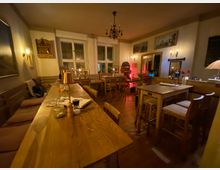 Gemütlicher Gastraum eines Restaurants mit mehreren Holztischen, Sitzbank und Holzstühlen auf Holzboden. Warmes Licht von Wandleuchten und einem Kronleuchter, dazu große Fenster, gerahmte Bilder an den Wänden und ein Spiegel.