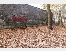 Ein leerer, mit Herbstlaub bedeckter Hof mit einer alten Backsteinmauer, auf der Graffiti zu sehen sind. Im Vordergrund stehen eine einfache Holzbank, ein Baum und ein verfallenes Holzspielgerüst mit Leiter.
