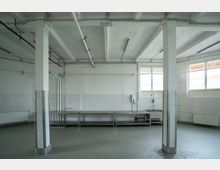 Industrieartiger Raum mit hellen Wänden, freigelegten Rohren an der Decke und mehreren Metalltischen entlang der Rückwand. Der Boden ist glatt und blassgrau, und große Fenster lassen natürliches Licht herein.