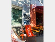 Street-side café exterior with an orange entrance wall and awning, a hanging sign reading “Jane and Jam,” and outdoor seating including wooden benches and branded deck chairs. A gray stucco wall with graffiti and a large window display sits beside small tables set with drinks, under dappled tree shade.