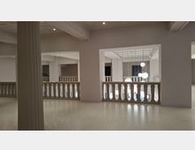Innenraum eines modernen Museums oder Foyers mit weißen Wänden, Balustraden und einem offenen Atrium. In der Mitte hängen mehrere kugelförmige Pendelleuchten, während Säulen und breite Deckenbalken die Architektur gliedern.
