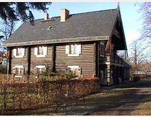 Holzhaus im traditionellen Stil mit dunklen Holzbalken und weiß gerahmten Fenstern; das Gebäude hat ein großes Satteldach und ist von einer Hecke und kahlen Bäumen in einer parkähnlichen Umgebung umgeben. Ein leerer Sitzbereich mit Bank befindet sich im Vordergrund.