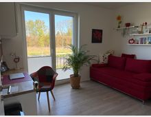 Heller Wohnraum mit großer Glasbalkontür und Blick ins Grüne, einem roten Sofa an der rechten Wand und einer großen Zimmerpflanze davor. Links steht ein kleiner Arbeitsbereich mit Holztisch und Stuhl; an der Wand hängen Regale mit Büchern und Dekoration.