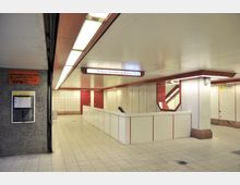 U-Bahn-Station mit hellen, gefliesten Wänden und roten Akzenten, einem Schild mit der Beschriftung „Rathaus Spandau ↔ Rudow (U7)“ und einem Rolltreppenbereich, der rechts nach oben führt. Links sind eine Infotafel und ein orangefarbenes Hinweisschild zu sehen.