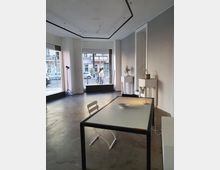 Ein minimalistisch eingerichteter Raum mit großen Glasfenstern, die den Blick auf eine belebte Straße freigeben. Im Vordergrund steht ein einfacher Tisch mit einem Stuhl, darauf befinden sich eine Schale und eine Lampe; im Hintergrund sind zwei Podeste mit dekorativen Objekten zu sehen.