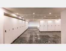 Ein heller Umkleideraum mit weißen Spinden auf beiden Seiten sowie grauem Marmorfußboden; die Decke ist mit kleinen eingebauten Leuchten versehen.