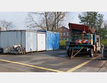 Ein Lagerplatz mit einem alten, rostigen Baumaschinenfahrzeug rechts im Bild, mehreren gestapelten Kunststoffteilen und Betonblöcken links sowie blauen und weißen Containern im Hintergrund. Im Hintergrund sind kahle Bäume und ein Backsteingebäude zu sehen.