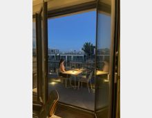 Eine Person sitzt bei Dämmerung auf einem modernen Balkon an einem Tisch mit einer kleinen Lampe. Im Hintergrund sind Gebäude und ein wolkenloser Himmel zu sehen, eingerahmt von geöffneten Glastüren.