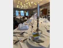 Ein elegant gedeckter Esstisch in einem Restaurant oder Veranstaltungsraum, mit weißer Tischdecke, Weingläsern, Besteck, einer brennenden weißen Kerze in einem silbernen Kerzenhalter und einer kleinen Schale mit Öl. Im Hintergrund sind verschwommene Lichter und weitere gedeckte Tische zu sehen.