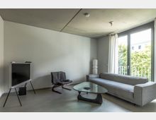 Wohnzimmer in modernem Stil mit grauem Sofa, Glastisch und einem schwarzen Stuhl mit Holzgestell. Große Fenster mit weißen Vorhängen bieten Ausblick auf grüne Bäume, und eine minimalistische Lampe steht in einer Ecke des Raumes.