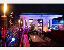Beleuchtete Dachterrasse bei Nacht mit Weihnachtsbaum, Holztisch und Stühlen. Im Hintergrund ein hell erleuchtetes Gebäude mit großen Fenstern sowie Fackeln und Lichterketten an der Brüstung.
