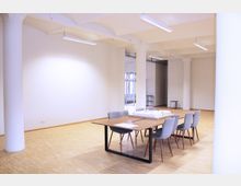 Großer, heller Raum mit hellem Holzboden, weißen Wänden und Säulen. In der Mitte des Raums steht ein rechteckiger Tisch mit grauen Stühlen, darauf liegt ein Architekturmodell.