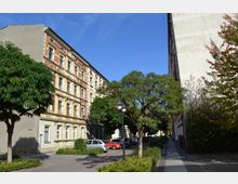 Straßenansicht mit mehrstöckigen Wohngebäuden im Altbaustil, flankiert von Bäumen und einem Gehweg. Im Vordergrund sind parkende Autos sowie eine Straßenlaterne zu sehen, umgeben von Büschen und Grünflächen.