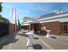 Außenbereich eines modernen Gebäudes mit roten Ziegelwänden und großen Fenstern, umgeben von einem gepflasterten Boden. Im Vordergrund stehen mehrere runde Stehtische mit weißen Hussen, daneben wehen zwei große Fahnen mit Schriftzügen.